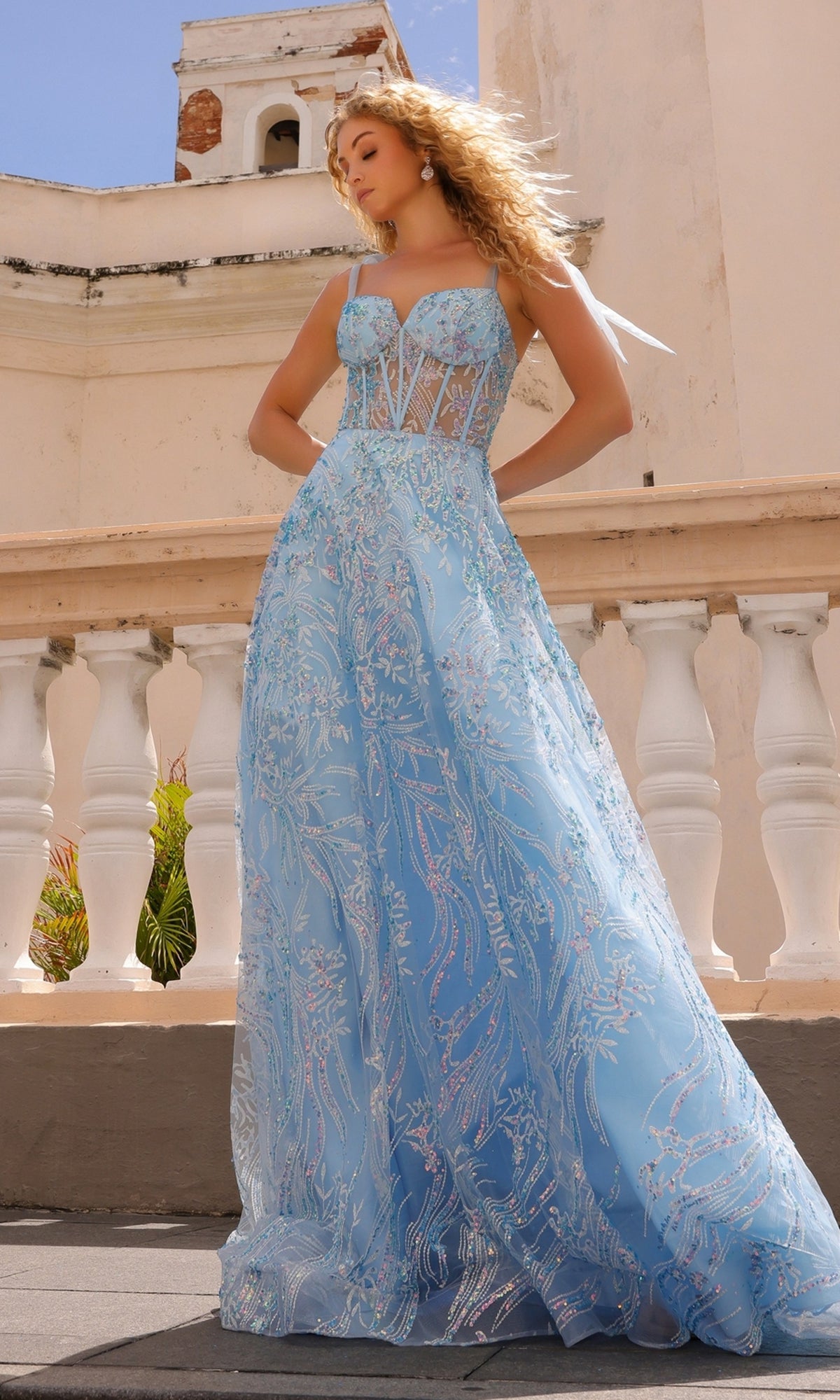 A woman stands outside a pale building in the Sequin Long Prom Ball Gown with Shoulder Bows T1336, a light blue, floor-length A-line dress featuring a corset-style bodice and silver embroidery.